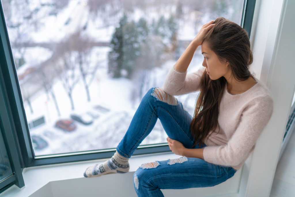 Woman with Seasonal Affective Disorder, or SAD, sitting on window ledge, looking at bad weather outside