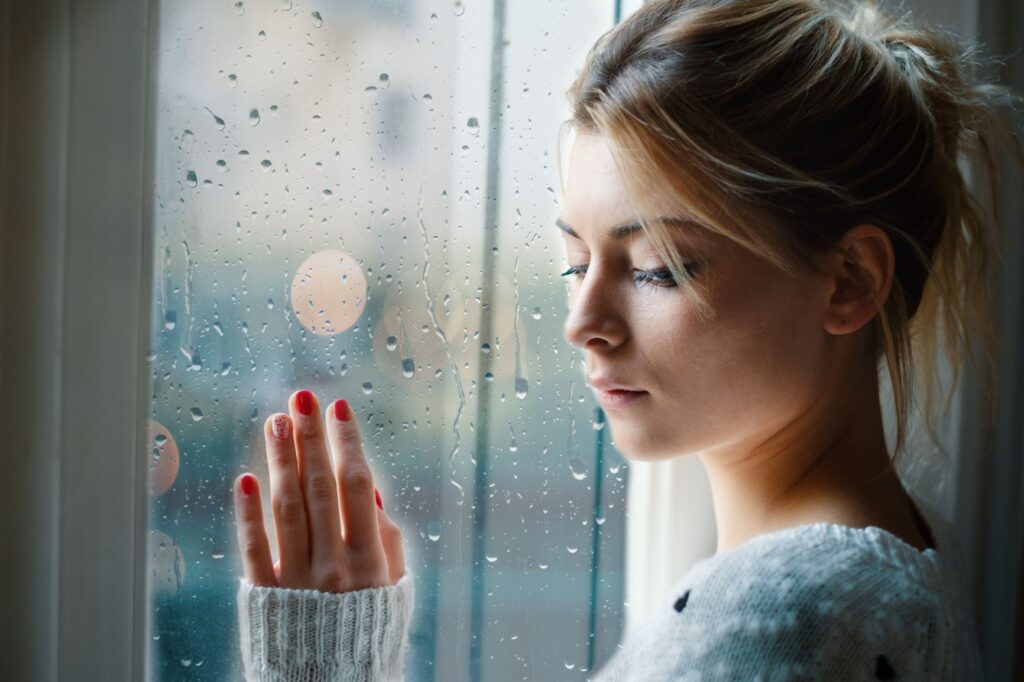 Side view of a sad girl leaning her left hand and right side of her forehead against a window on a rainy day.